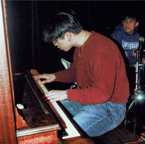Man in red sweater sitting at acoustic piano playing looking down at the keys.
