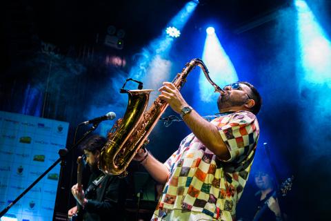 Diego Herrera playing a saxophone on stage under blue lights, with other musicians performing in the background.