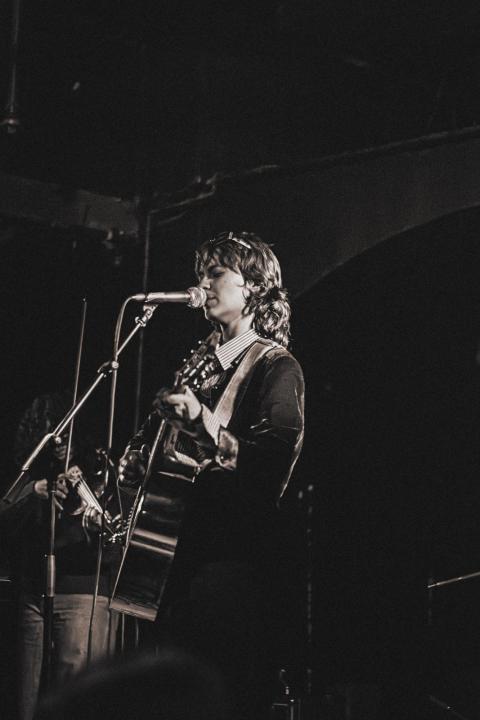     Black-and-white photo of Marianthi Olympia singing into a microphone while playing guitar on stage, with another musician partially visible in the background.