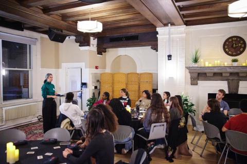 An indoor group presentation taking place in a room featuring coffered wooden ceilings and a large fireplace decorated with a clock and small plants. On the left, a female-presenting speaker wearing a teal turtleneck and black trousers stands facing the room, addressing an audience seated at round tables with dark tablecloths.