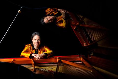 Kris Davis sits at an open grand piano. She is white with brown hair and wears a bright orange silk jacket
