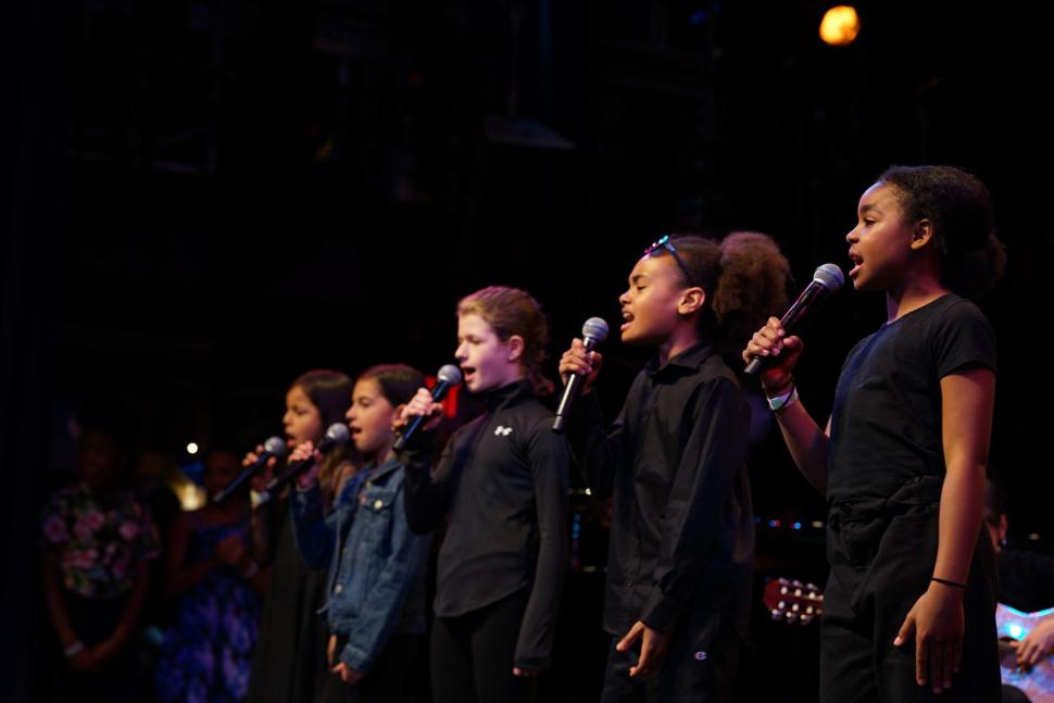 Five young students stand on stage, each singing passionately into individual microphones while wearing black tops and dark bottoms.